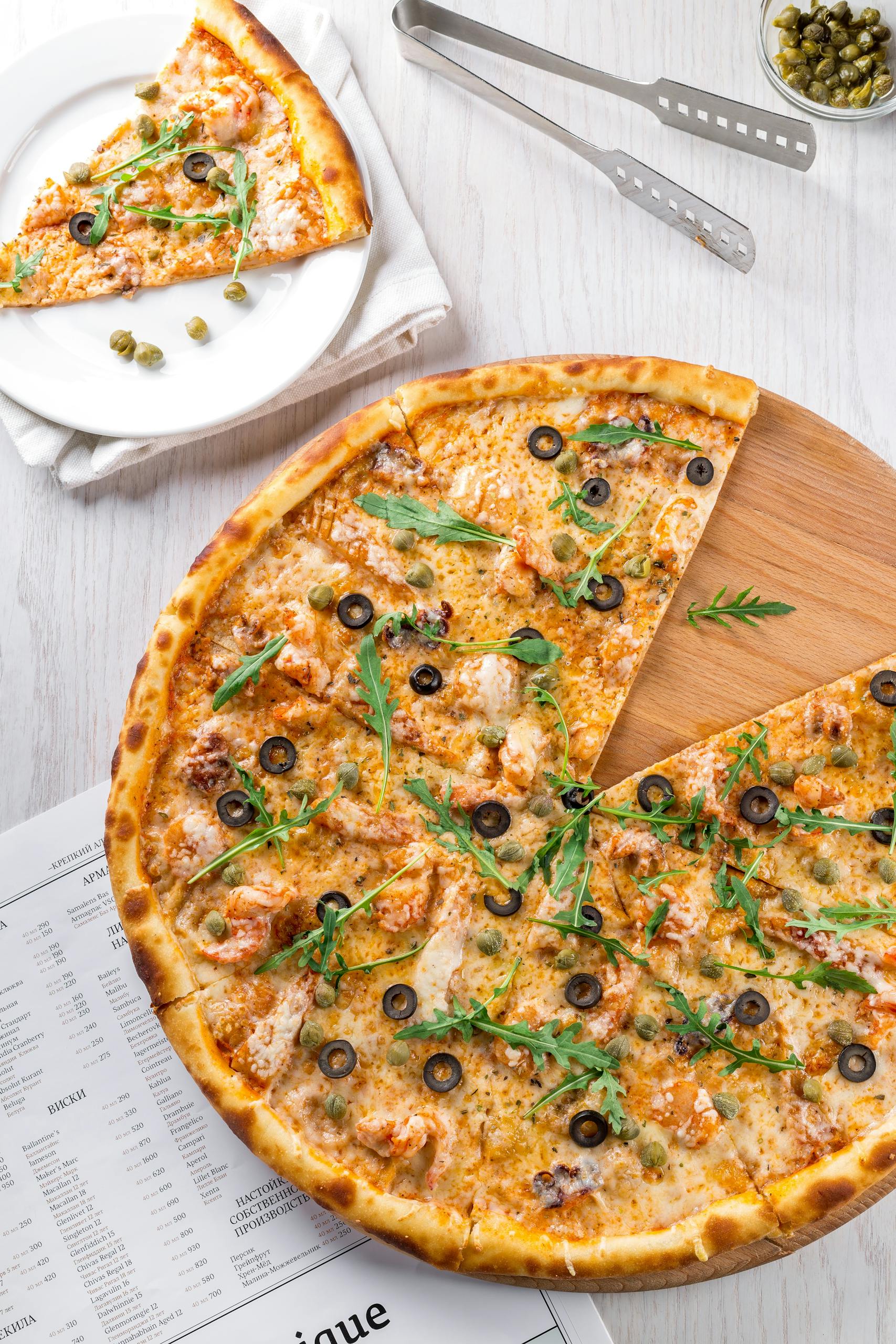 High-angle view of pizza topped with olives, arugula, and cheese. A mouthwatering slice on a plate nearby.